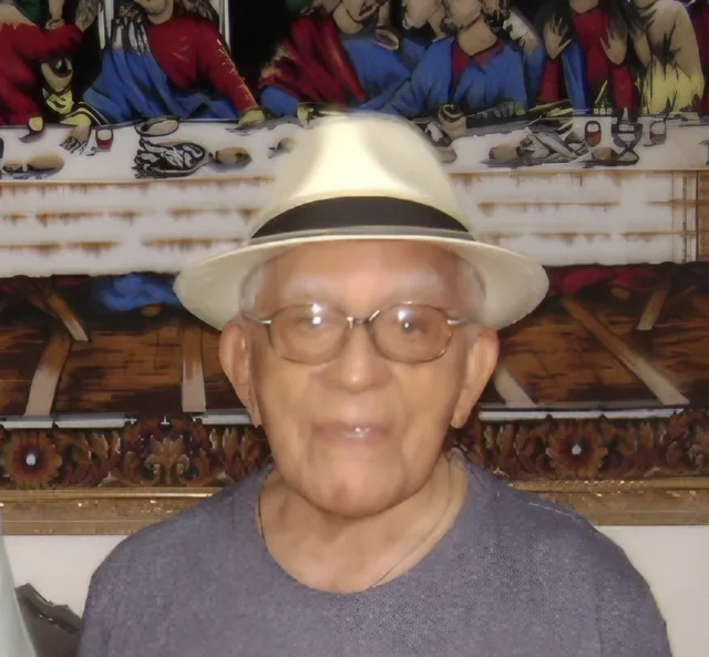 Retrato de Expedito, de chapéu claro e óculos, diante de uma tapeçaria da Santa Ceia